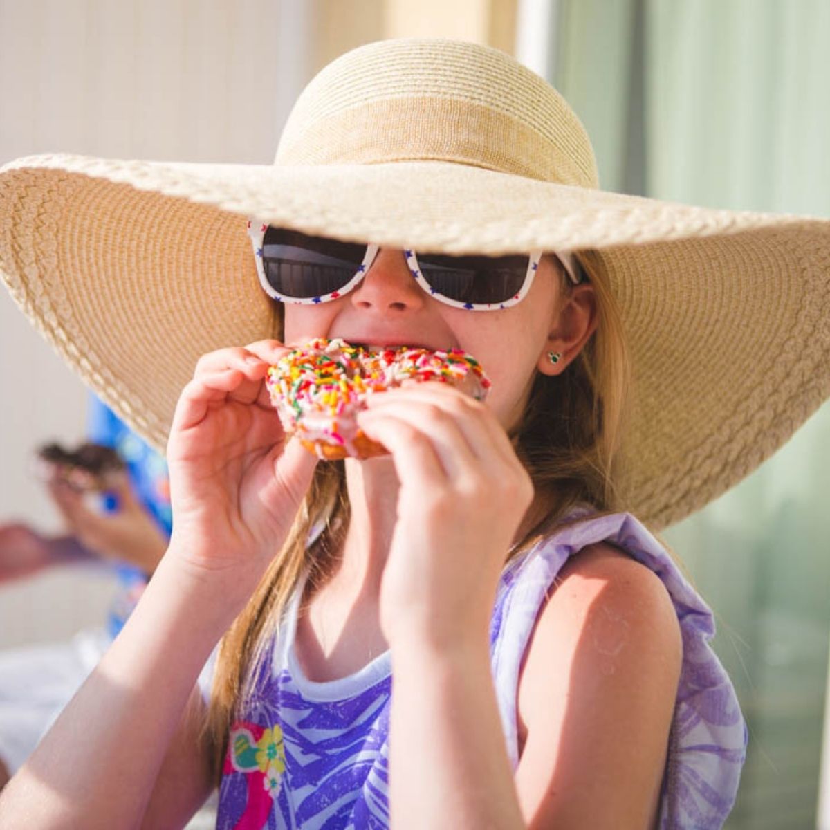 A young girl is eating a donut while wearing a sunhat and sunglasses.