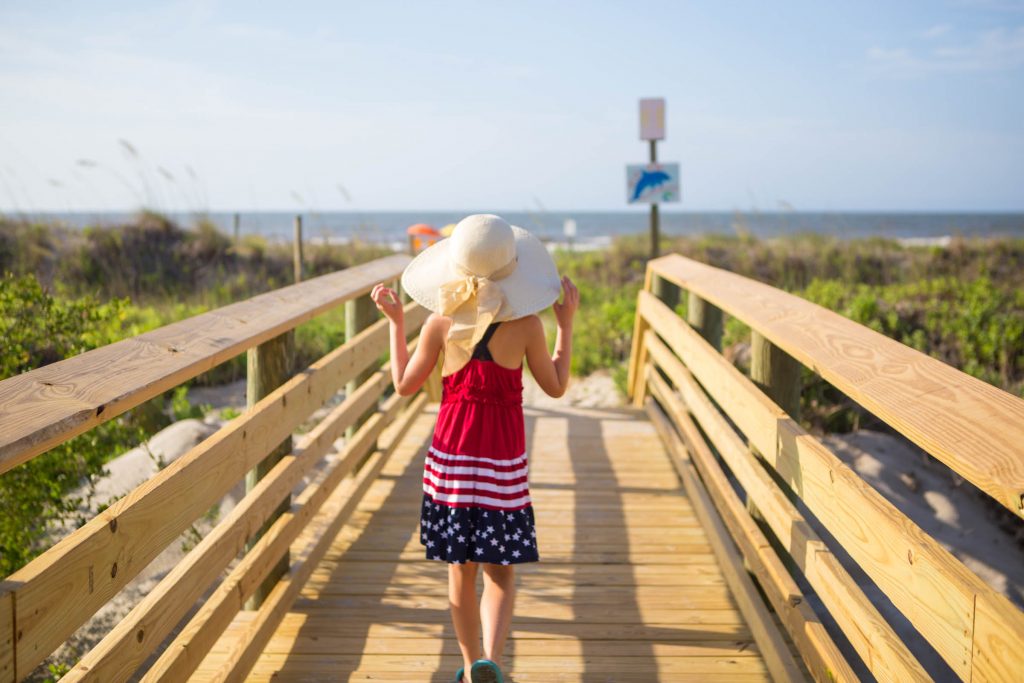 A young girl wearing a large sun hat walks on the pier towards the beach.