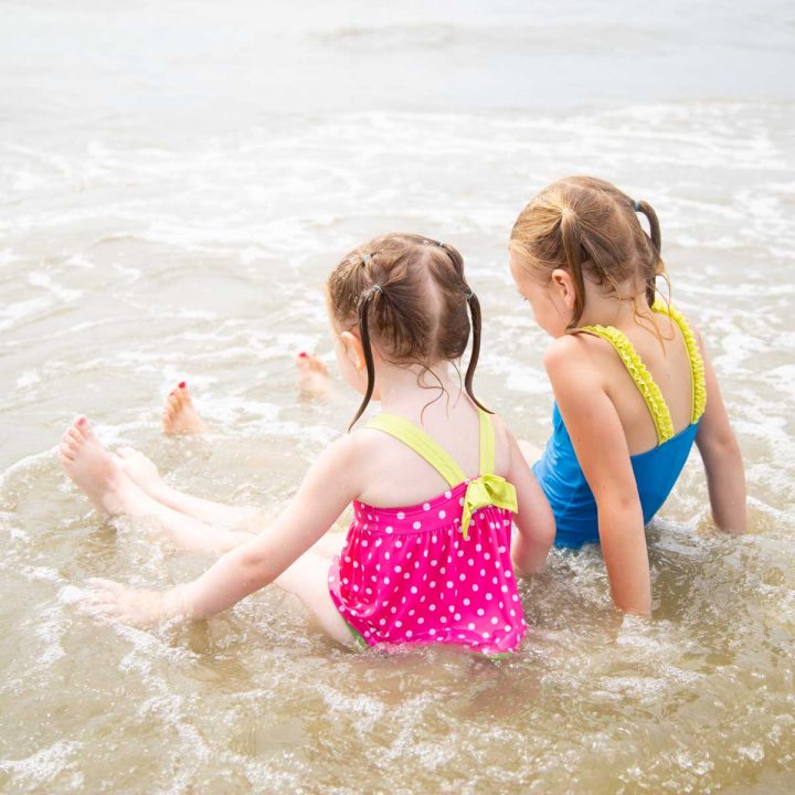 Two young girls sit in the shallow ocean water at the beach.