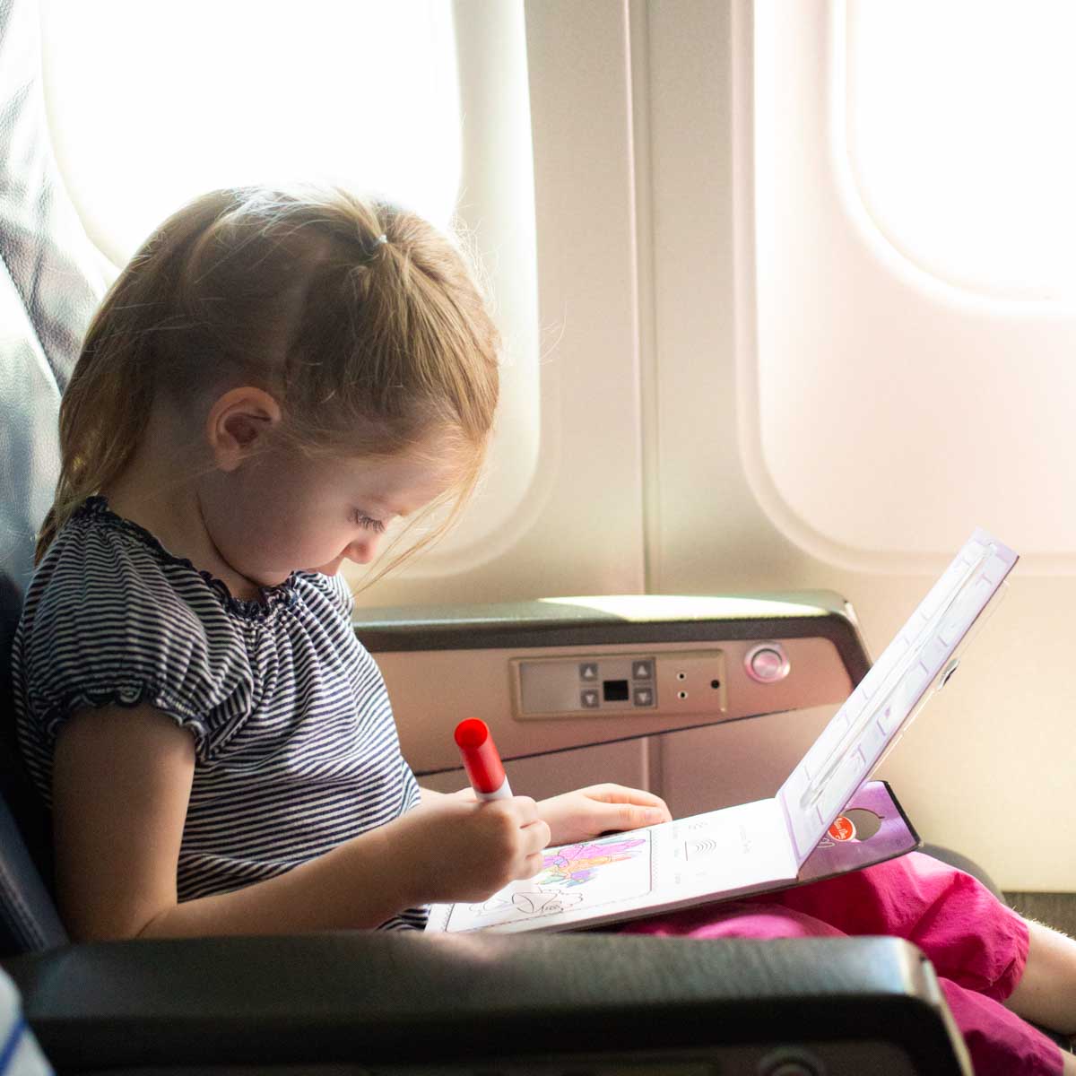 A young girl sits on an airplane and is coloring in her seat.