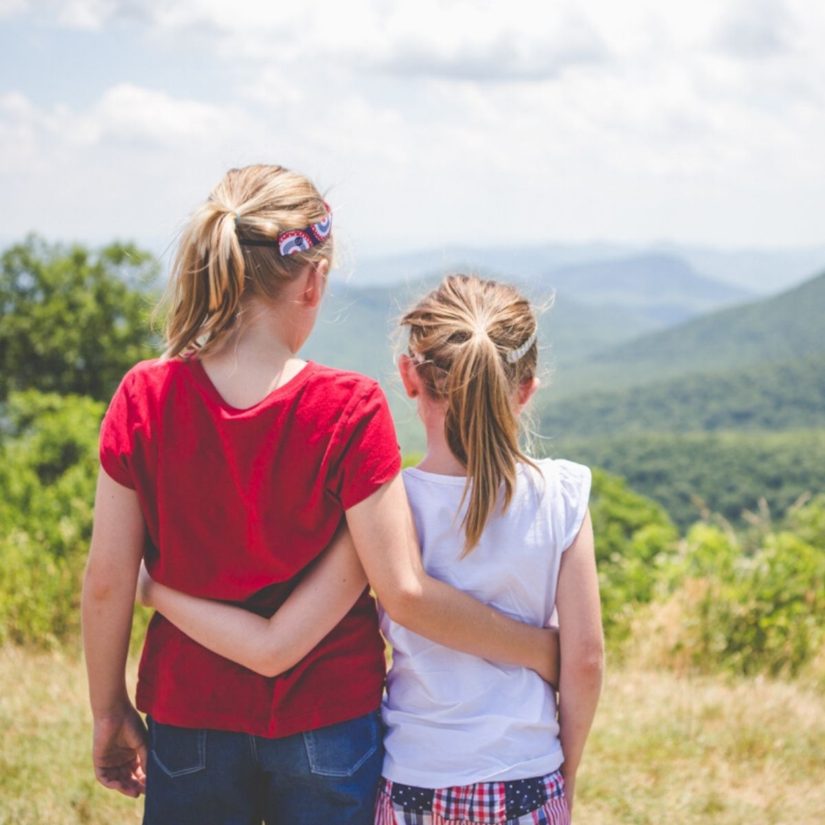 Two young girls have their arms around each other while looking out at the mountains.
