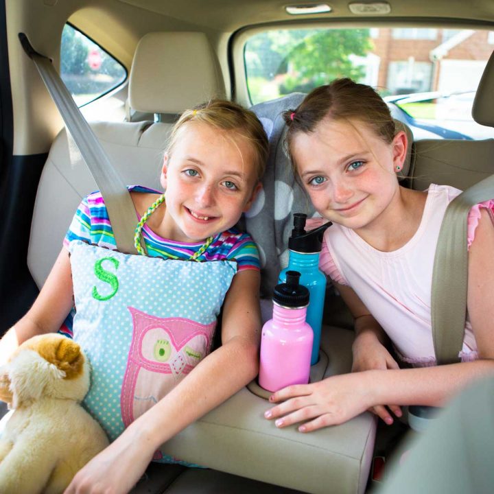 Two young girls in the back seat of a minivan are buckled in and ready for the road trip.