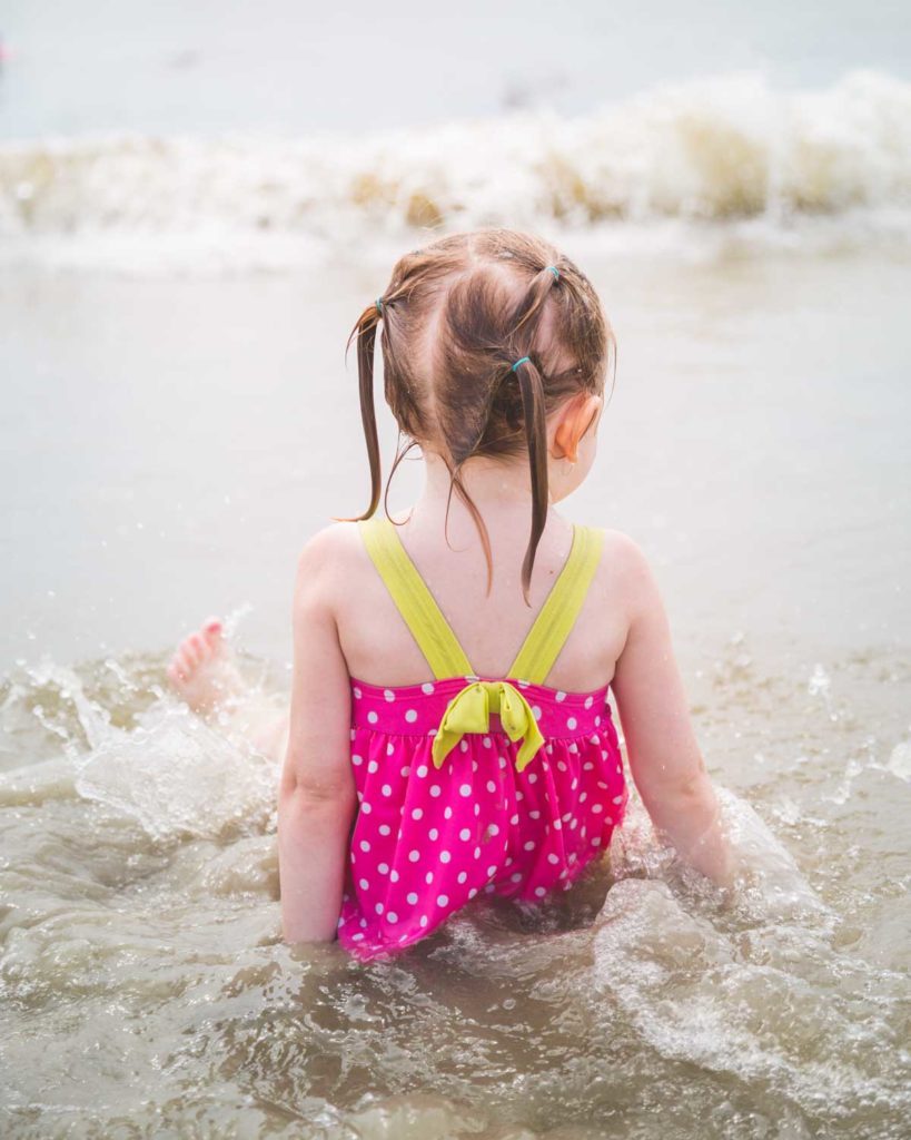 A little girl in a hot pink swim suit sits in the ocean water at the beach.