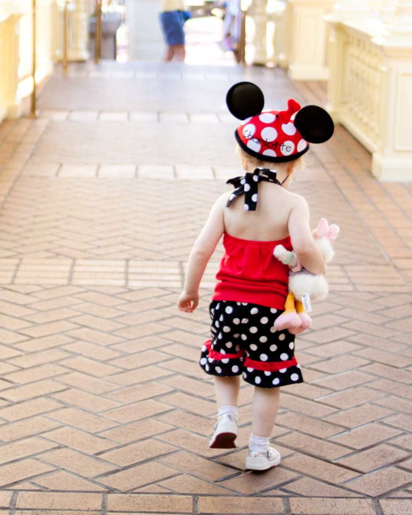 A toddler wearing a polka dot Mickey Mouse hat is walking at Disney World.