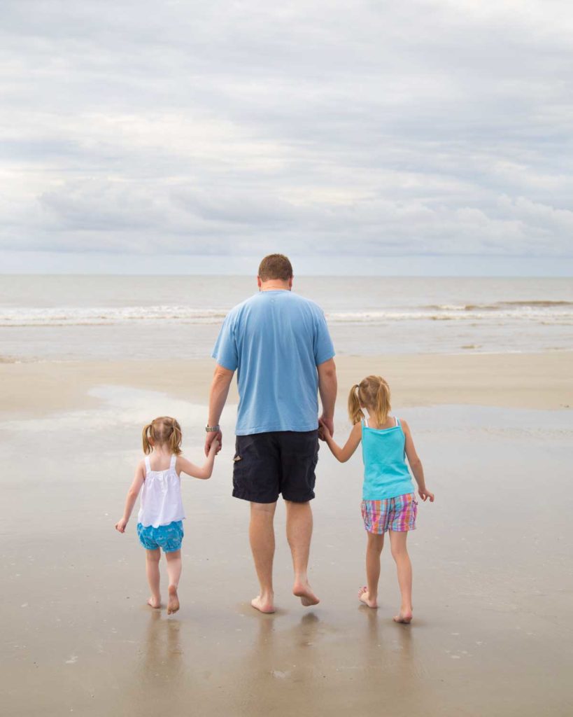 A dad holds the hands of his two daughters as they visit the beach for the first time.