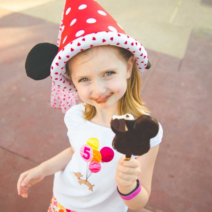 A young girl in a Minnie Mouse princess hat has taken a bite out of a Mickey pop and has chocolate on her face.