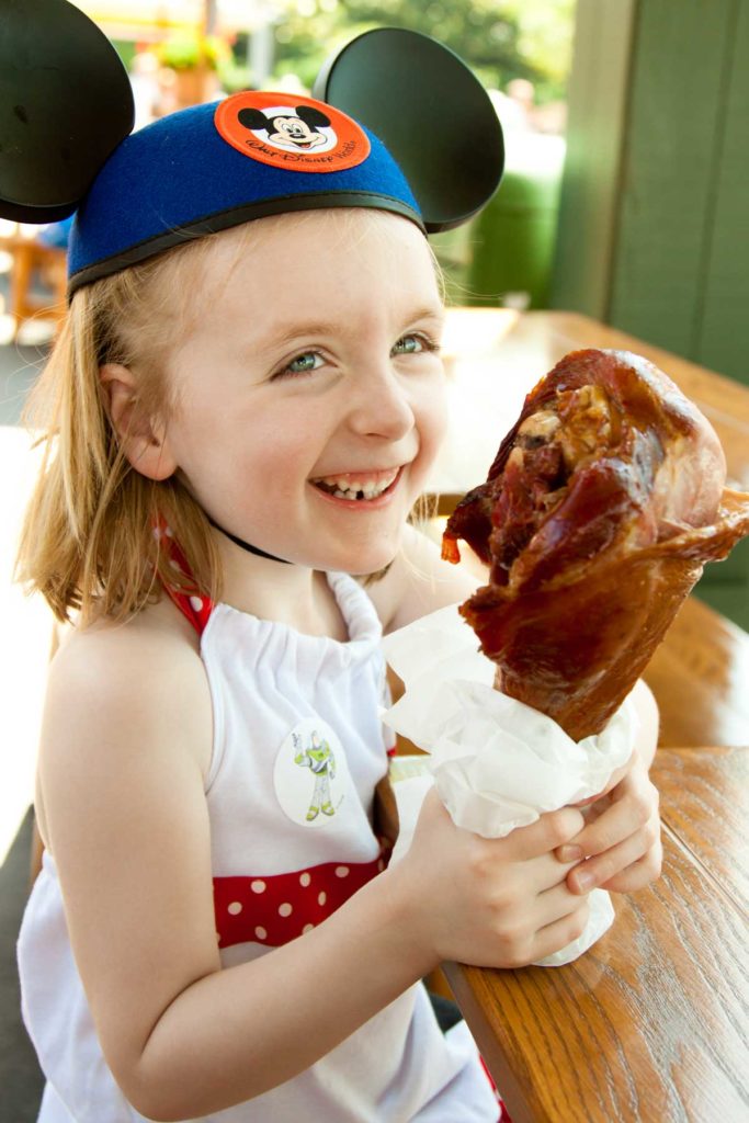 A young girl eats a giant turkey leg at Disney World.