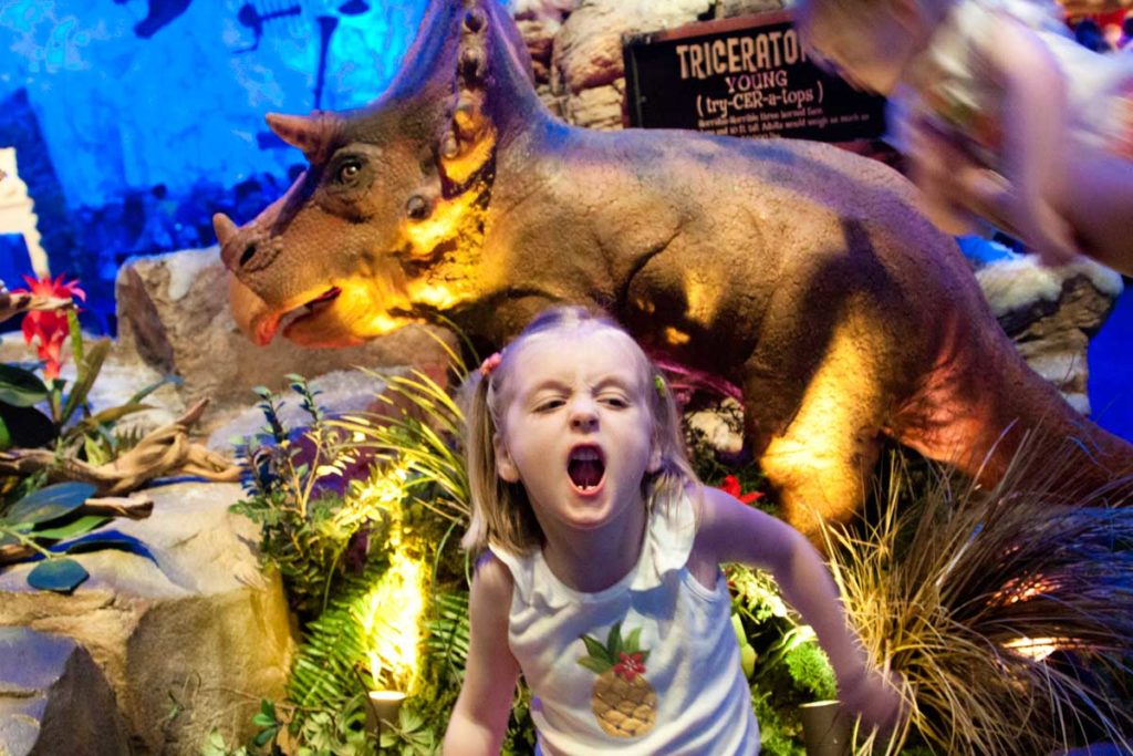 A young girl roars by a dinosaur at a restaurant.