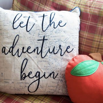 A pillow that says "Let the Adventure Begin" sits next to a red apple pillow.