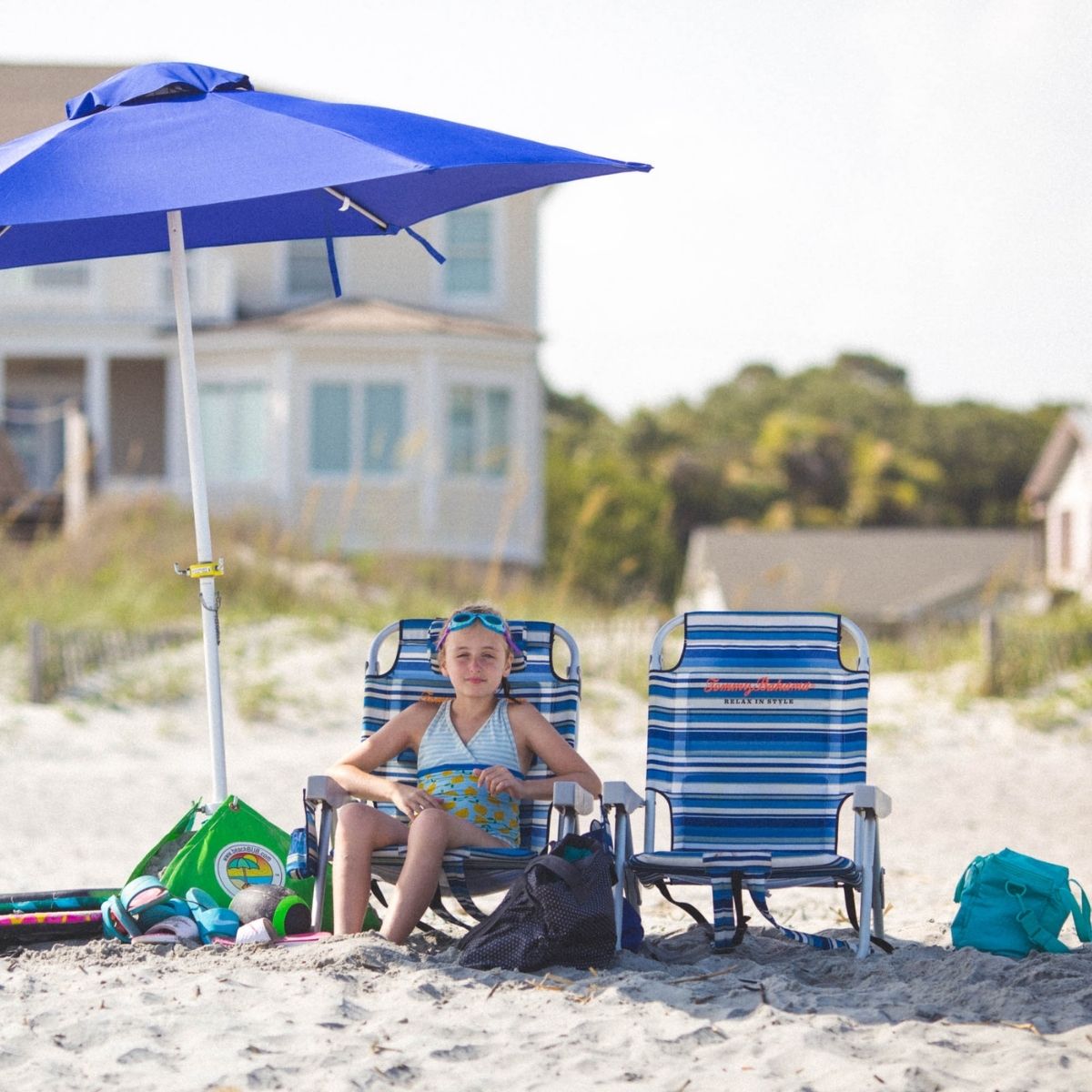A young girl sits on a beach chair under a beach umbrella.