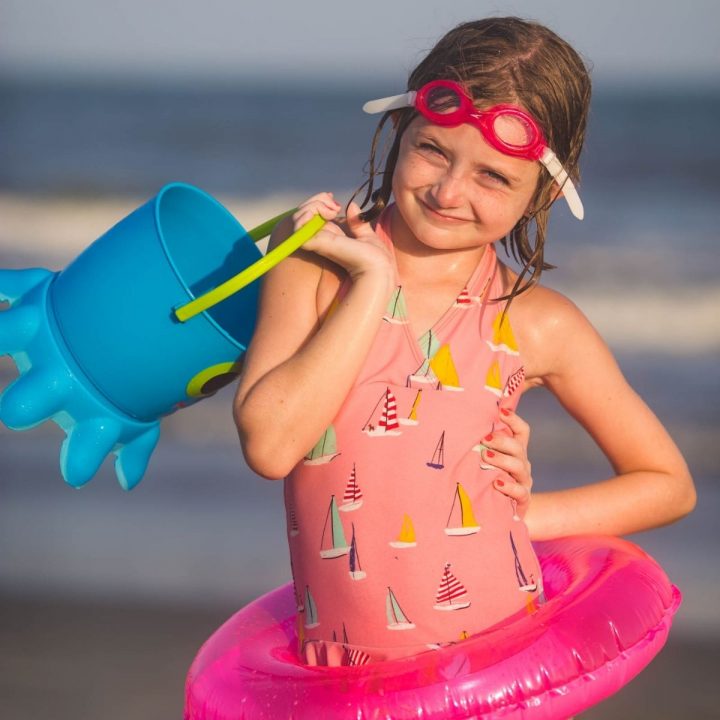 A young girl is posing with a beach pail and floatie with goggles on her head.