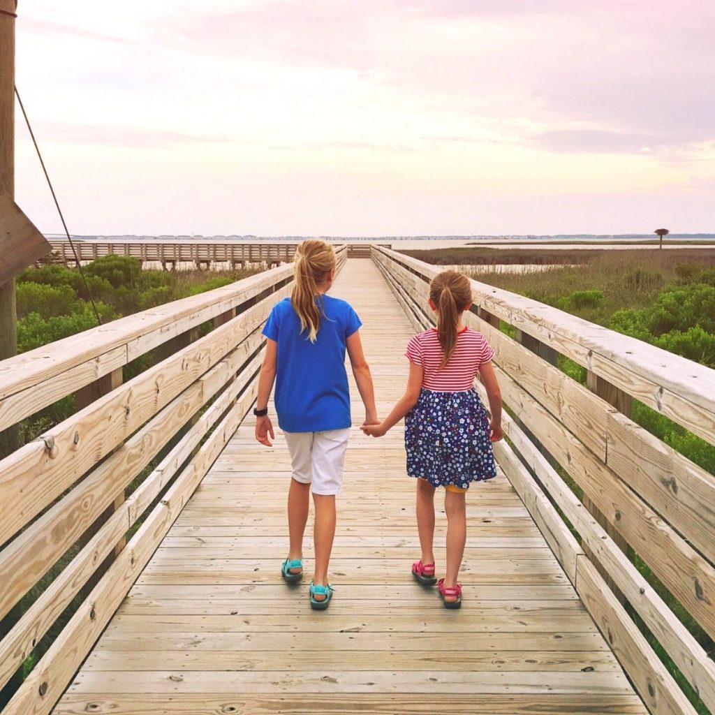 Two young girls holding hands walk down a beach pier.