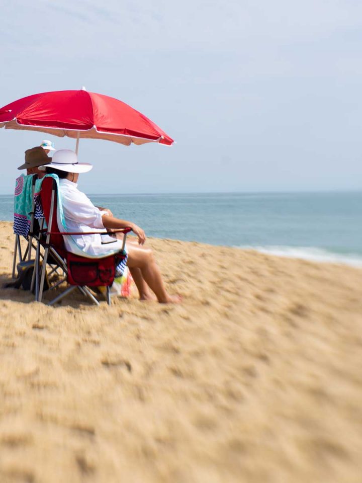 A couple sits underneath a red umbrella on the beach.