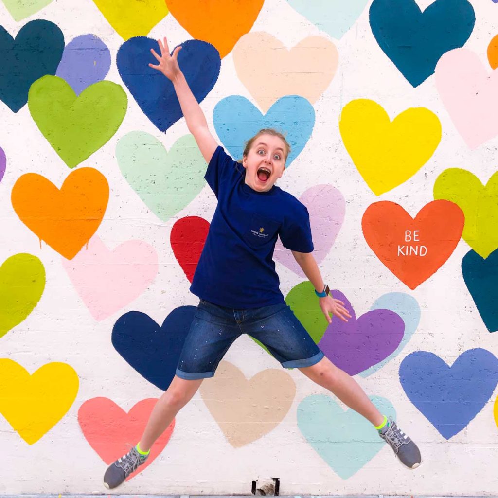 A young girl leaps in front of a painted mural that shows rainbow hearts against a white backdrop.