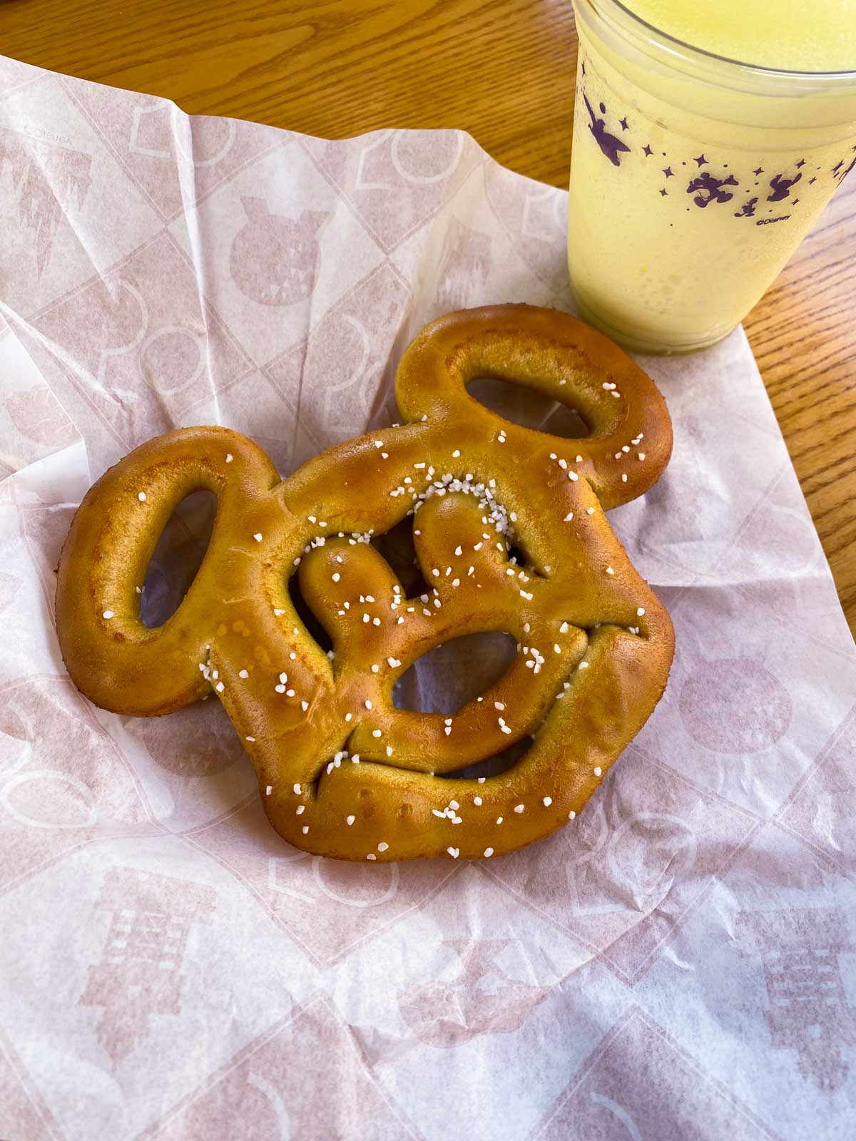 A salted Mickey Mouse shaped soft pretzel on a paper next to a frozen lemonade.