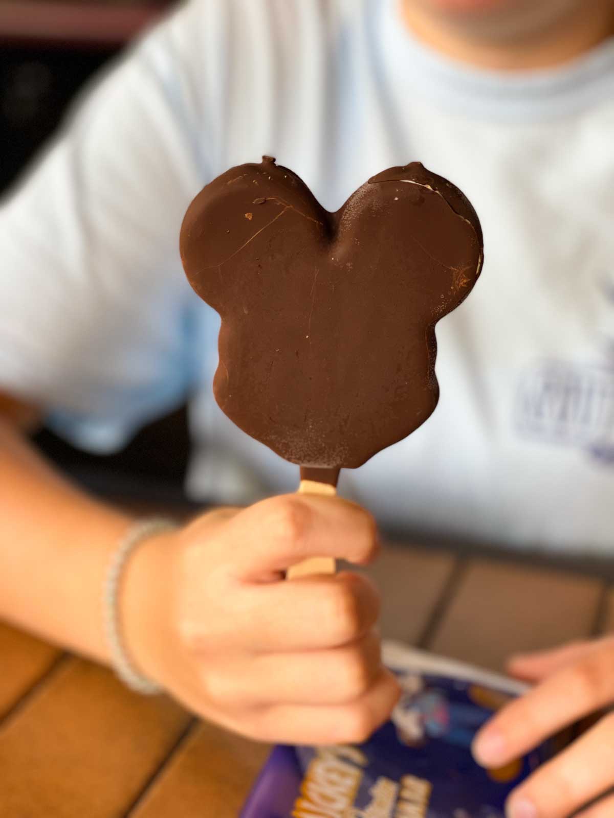 A mouse shaped chocolate covered ice cream pop is held by a girl.