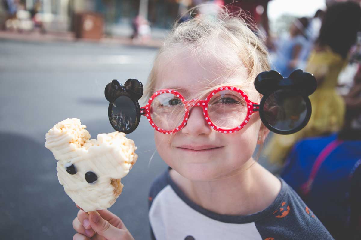 A young girl wearing Mickey Mouse glasses holds a Mickey Mouse rice crispy treat.