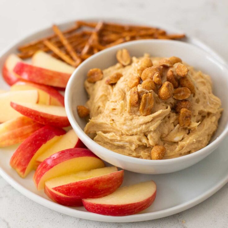 A plate of apple slices next to a bowl of peanut butter dip with chopped peanuts on top.