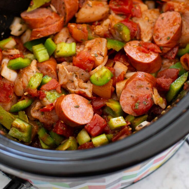 A slowcooker pot has chopped vegetables, sausage, and chicken.