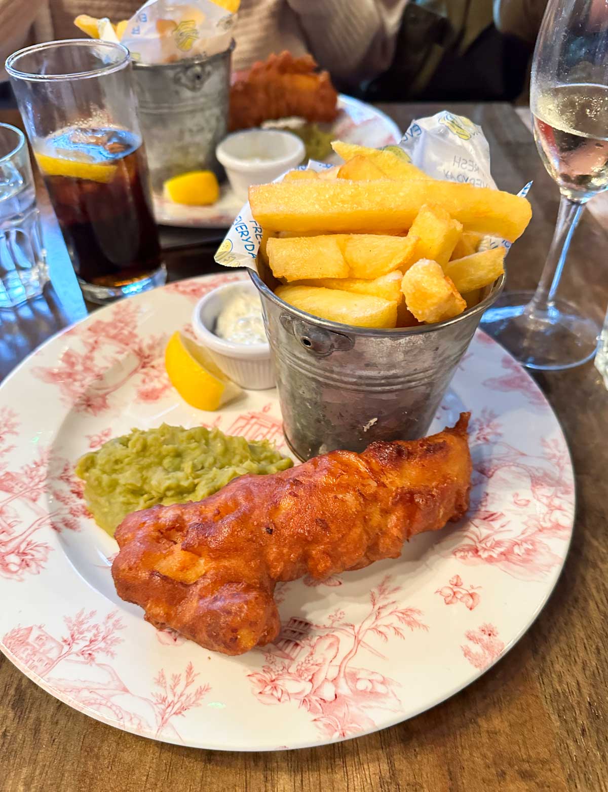 A plate has a piece of fried fish next to mushy peas and a container of chips/french fries.