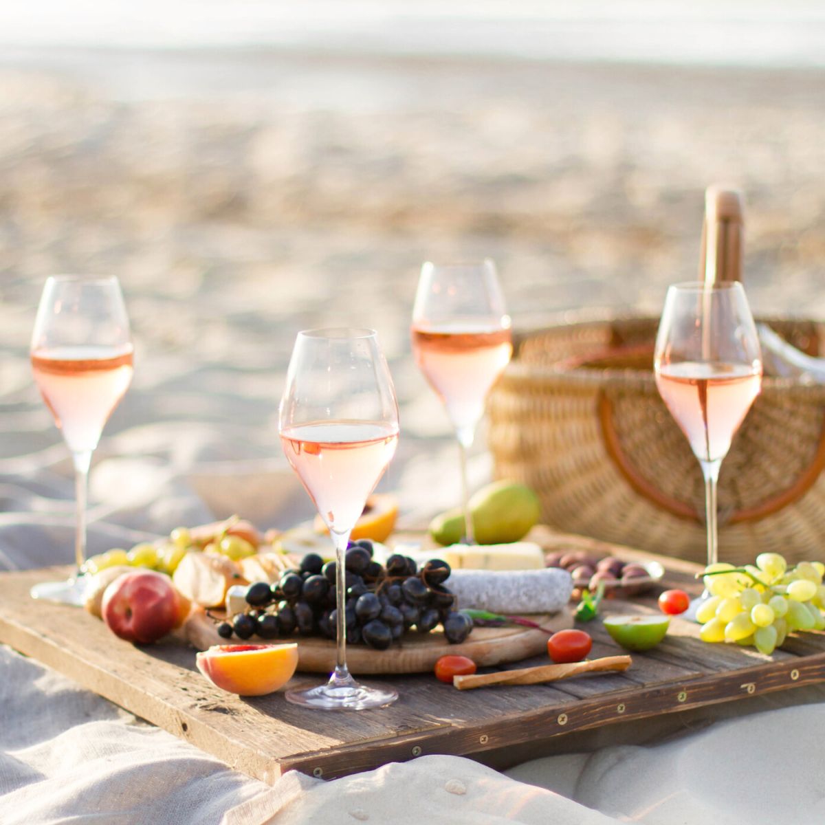 4 wine glasses and some fresh fruit and cheese sit on a wooden table on the sand next to a picnic basket.
