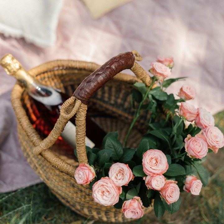 A wicker basket filled with pink roses has a bottle of rose tucked inside and sits on a light pink picnic blanket.