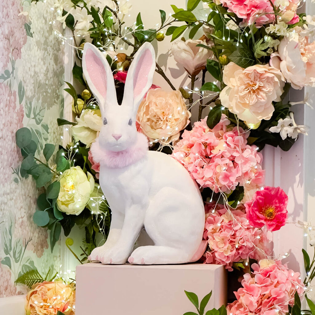 A white bunny statue sits on a pedestal decorated with pink flowers.