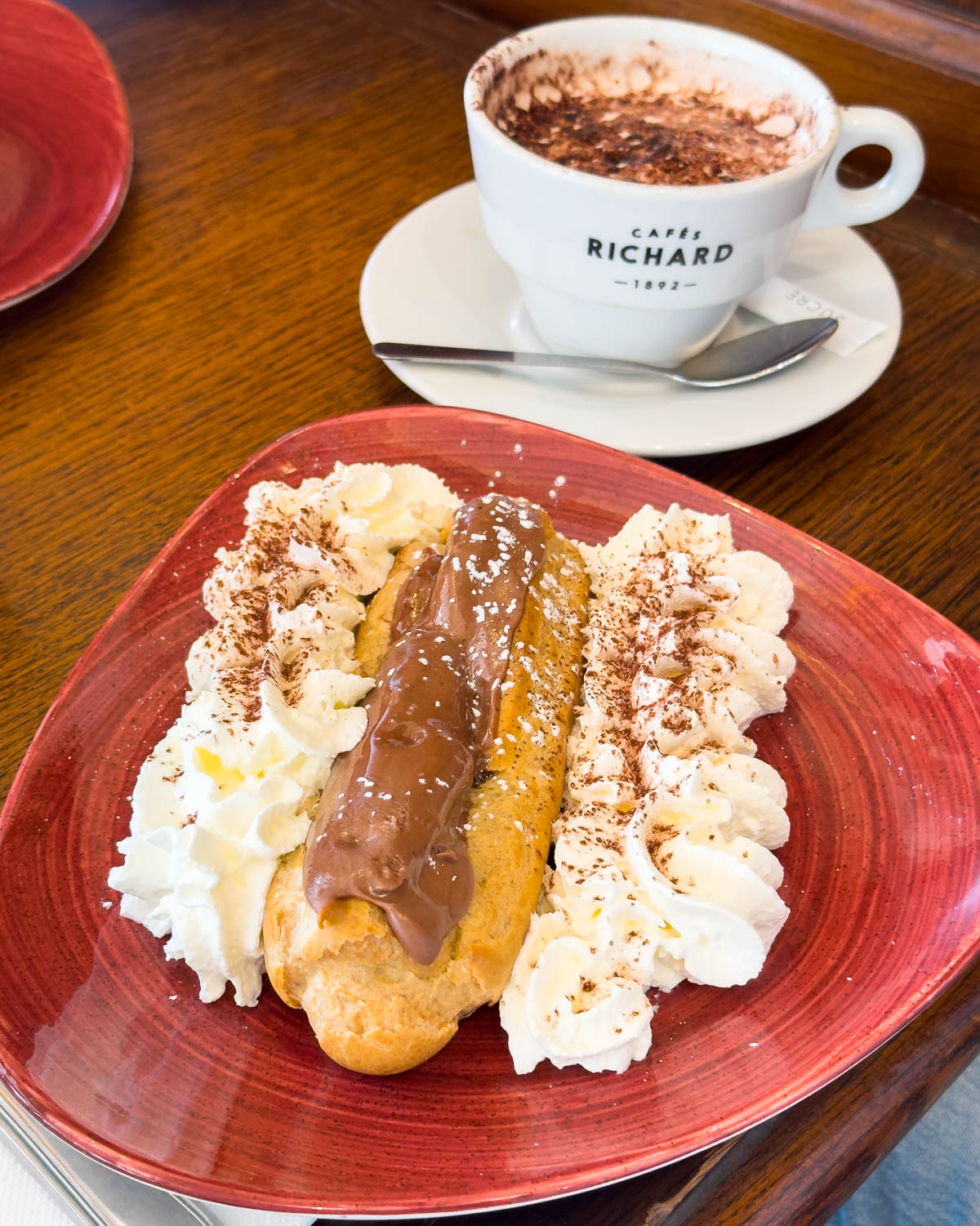 A plate with an eclair in the middle of two strips of whipped cream. A mug of hot cocoa on the side.