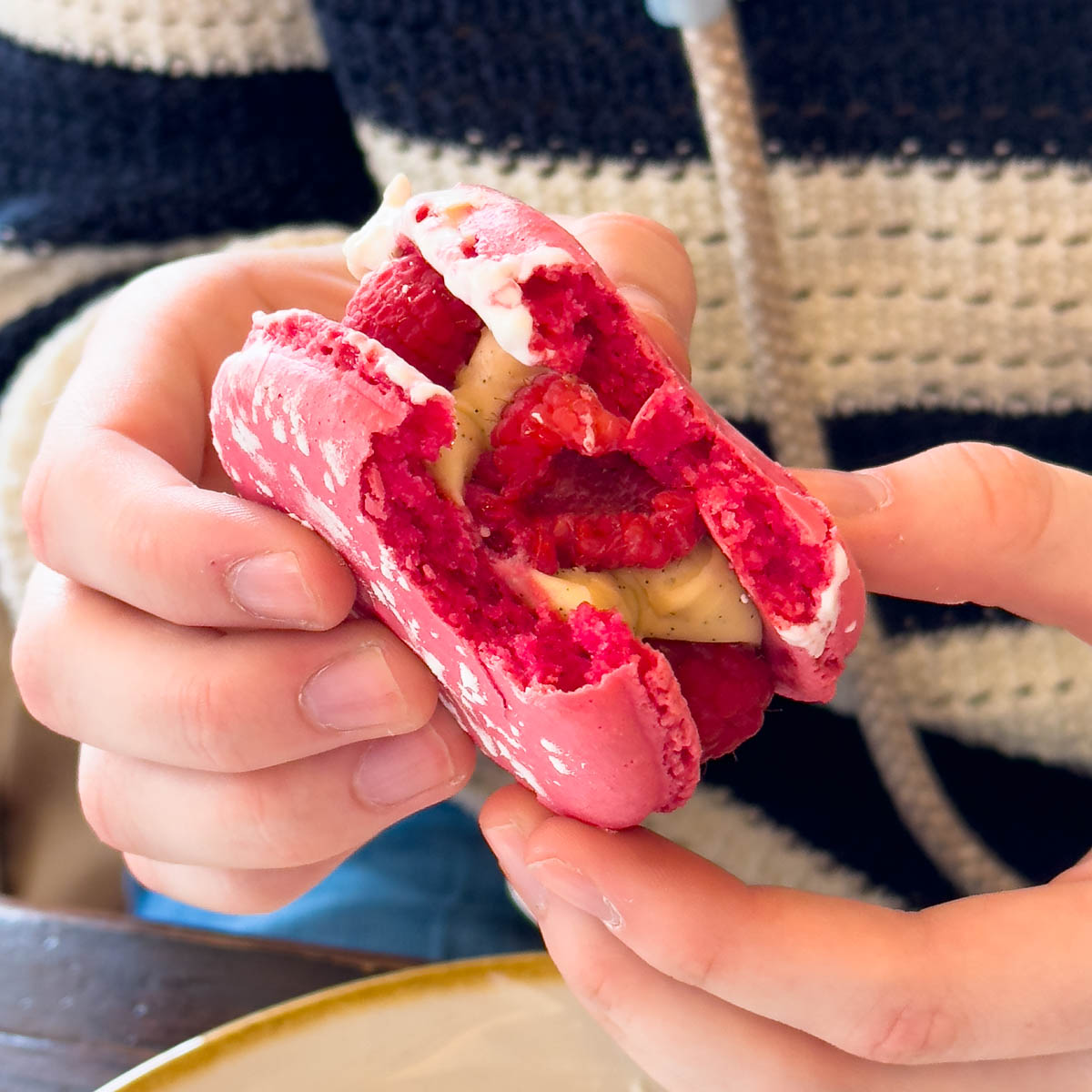 A girl holds a pink raspberry sandwich cookie with creamy filling and a fresh raspberry in the middle.