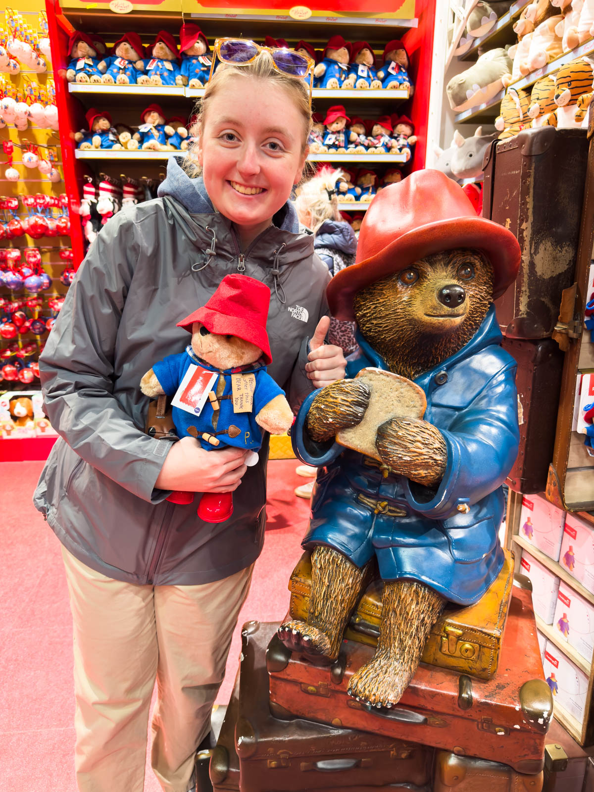 A young girl poses with a Paddington bear toy next to a Paddington bear statue.