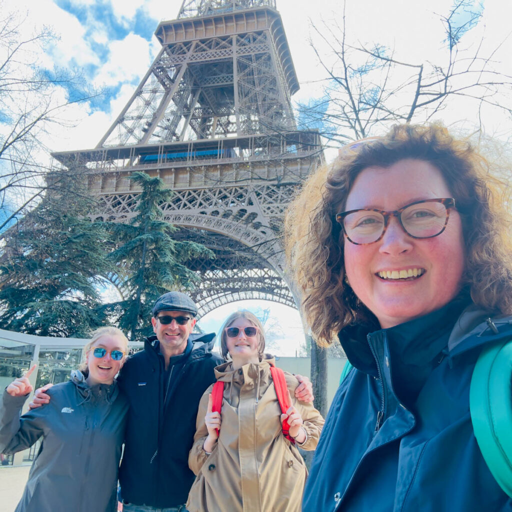 A mom takes a selfie in front of the Eiffel Tower in Paris. Her family stands in the back and are pointing up.