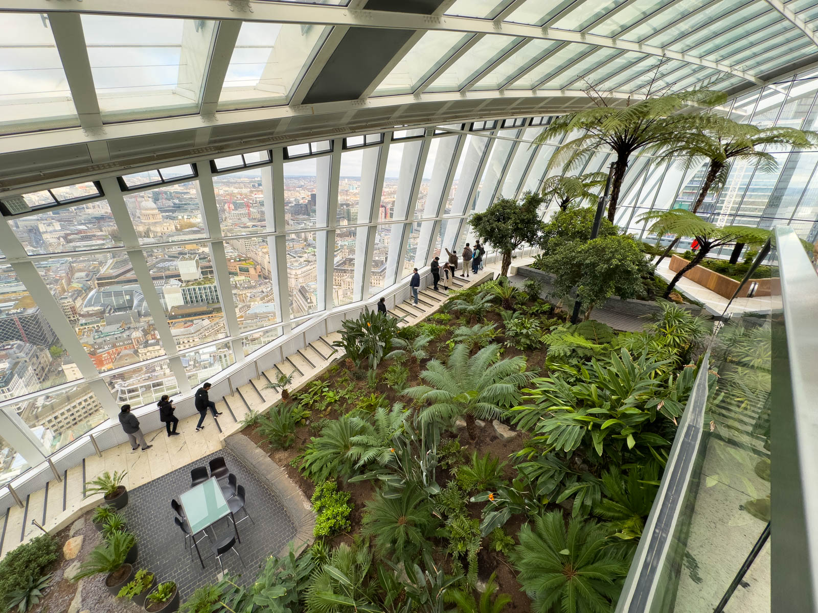 Tourists walking along the indoor garden near a wall of windows that look out at London.