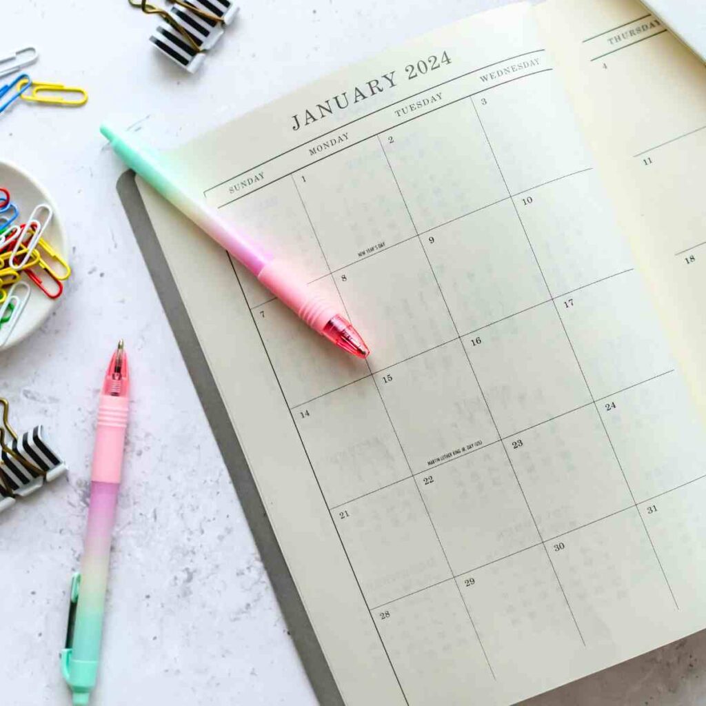 A calendar is on the desk with colorful pens around it.