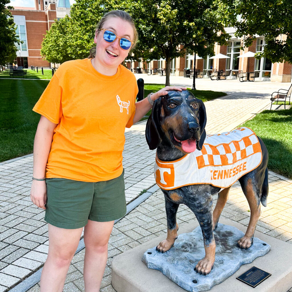 A prospective student wearing an orange UT t-shirt stands next to a dog statue on campus.