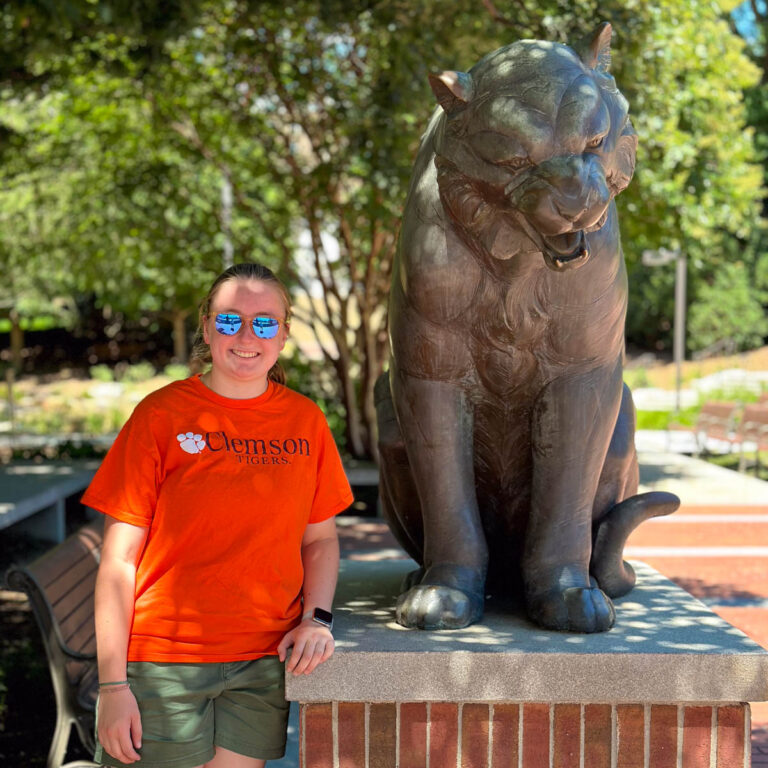 A student wearing an orange Clemson t-shirt stands next to a statue of the tiger mascot.