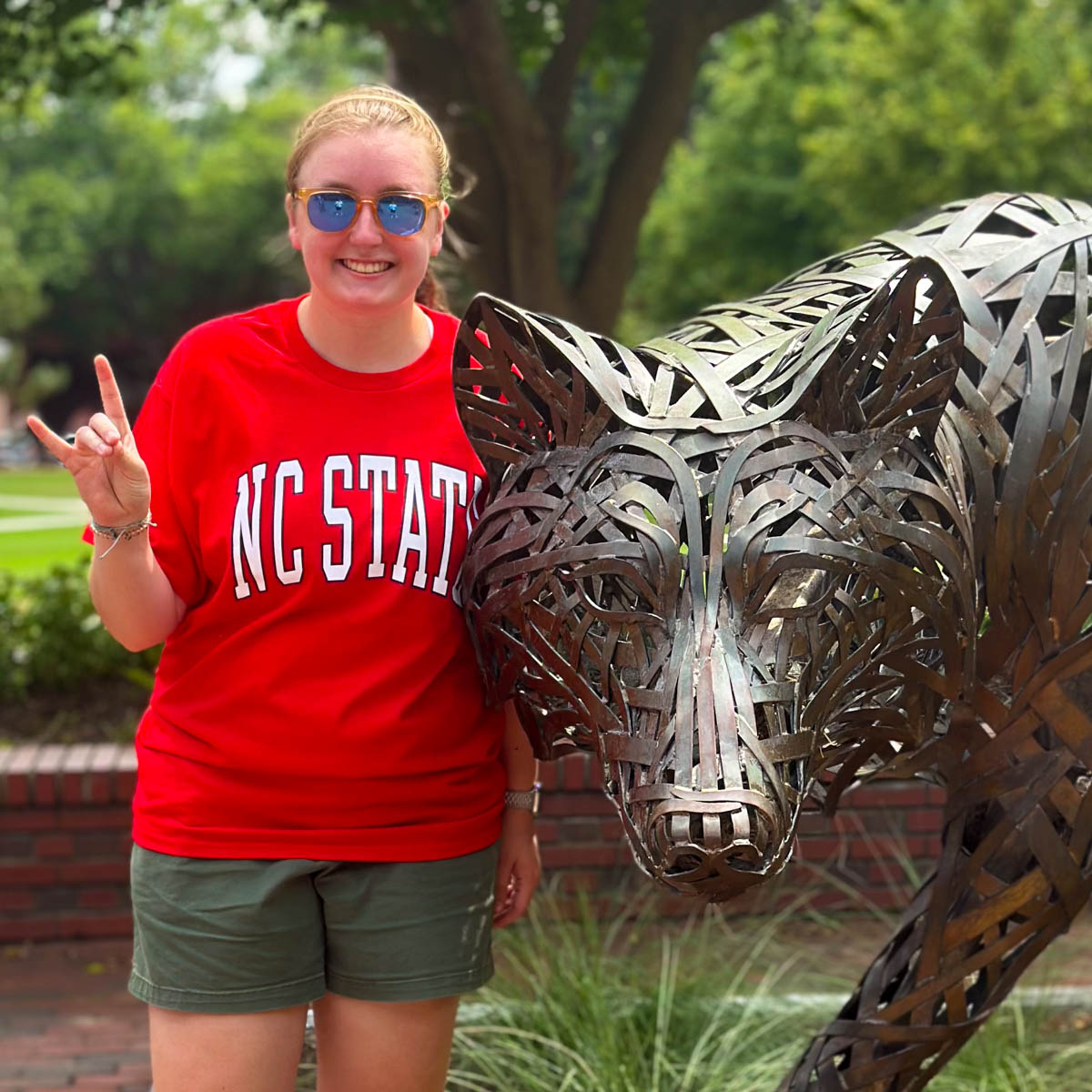 A girl wearing a red NC State t-shirt stands next to a statue of a wolf on campus.