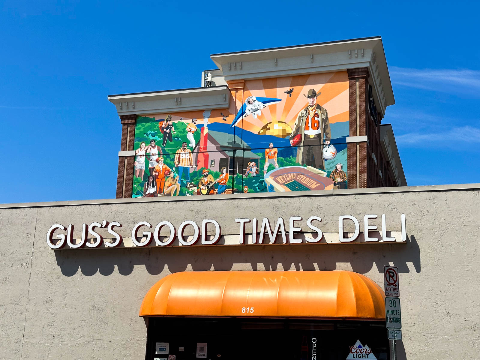 The outdoor sign of the deli in front, you can see a large Tennessee mural on the building just behind it.