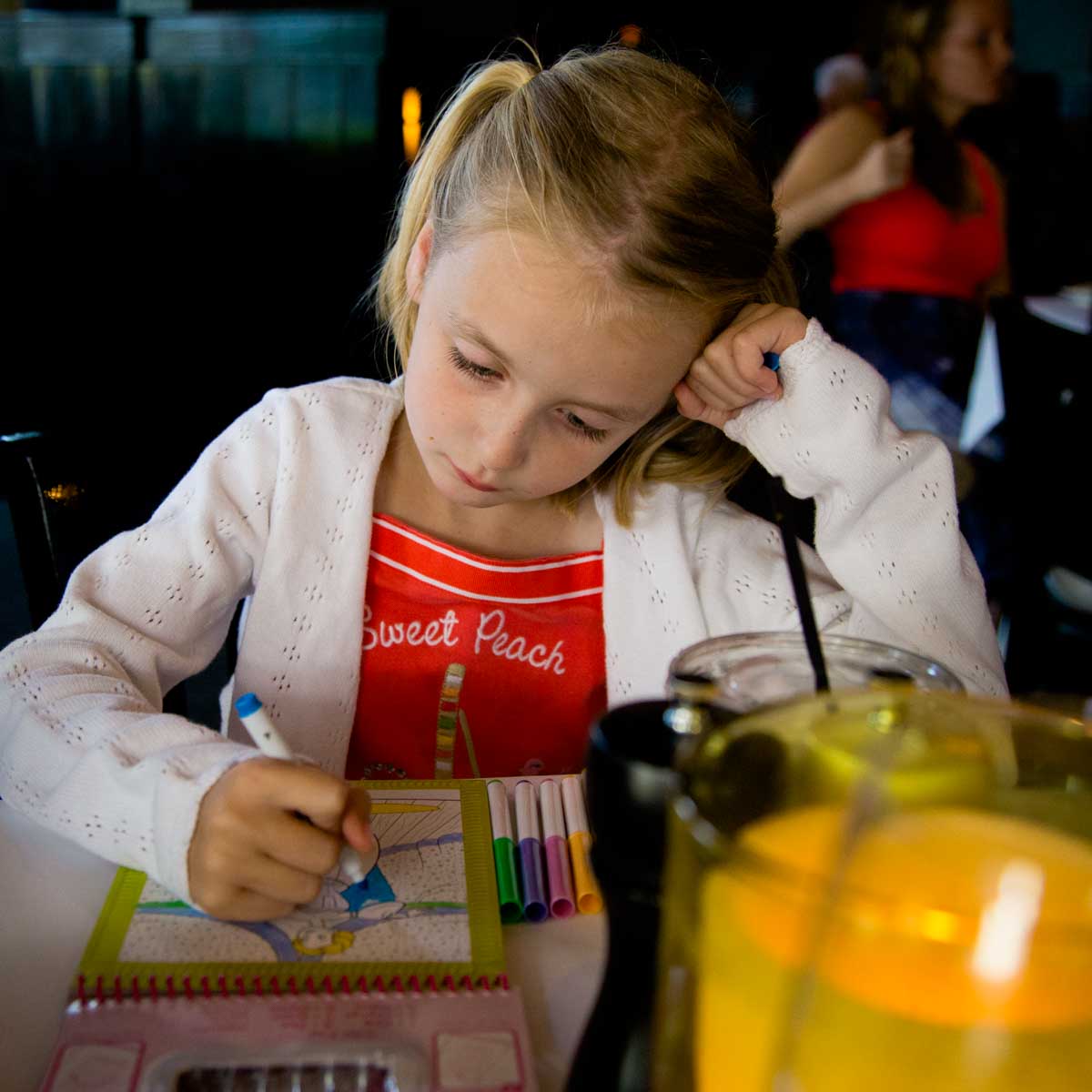 A young girl dressed up for a nice dinner in a restaurant is coloring quietly at the table.