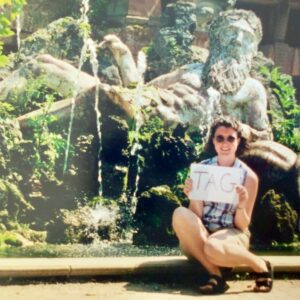 A vintage photo of a woman sitting in front of a fountain holding a sign that says "Tag"