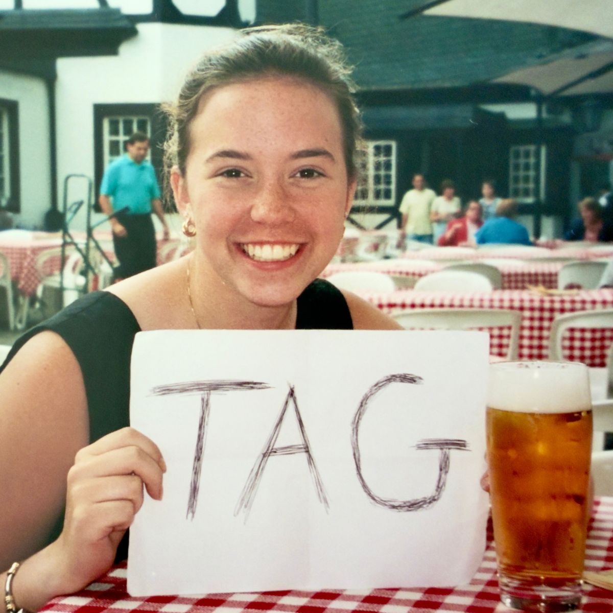A vintage photo of a young woman sitting in a German biergarten holding a sign that says "Tag"