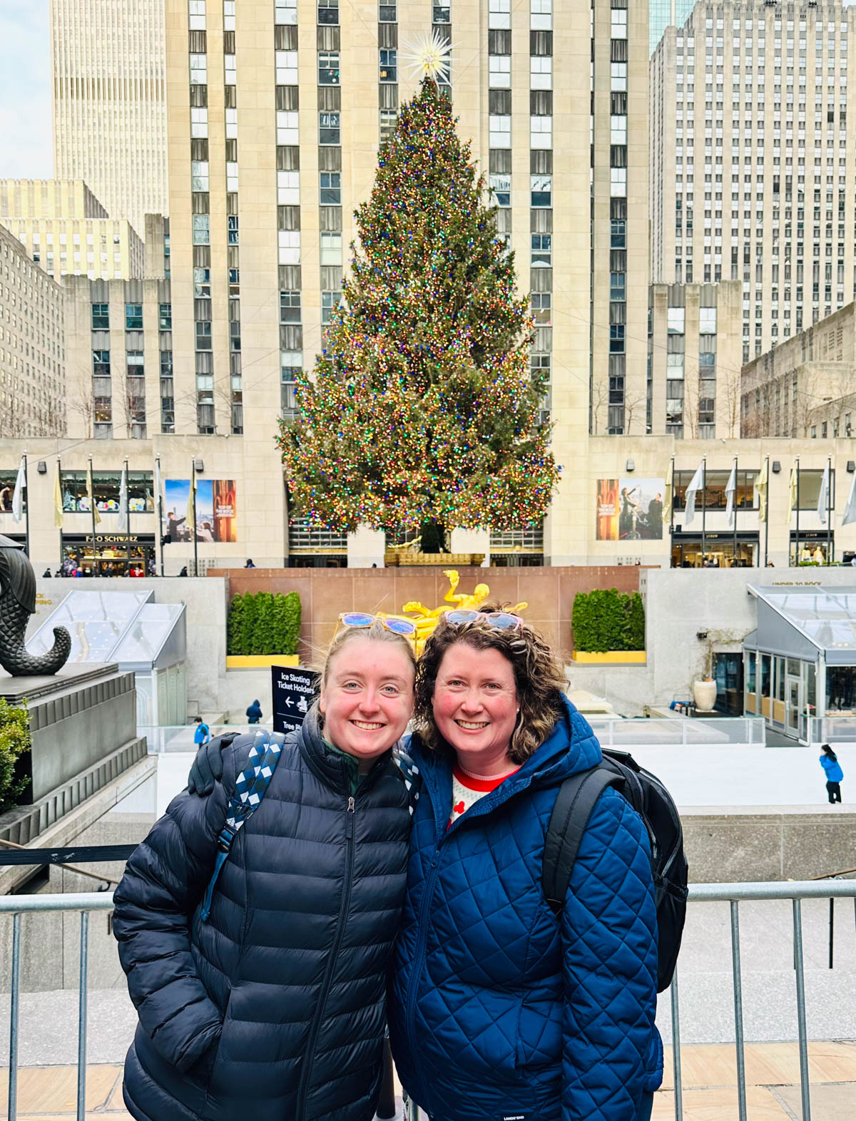 A mom and daughter pose in front of the Rockefeller Center Christmas tree in New York City at Christmas time. 